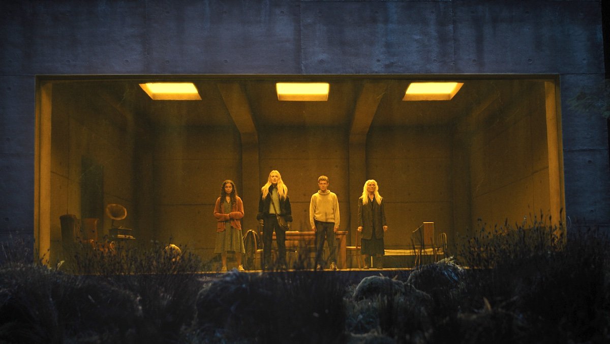 Four people stand in a well lit room at night in front of a giant window in The Watchers